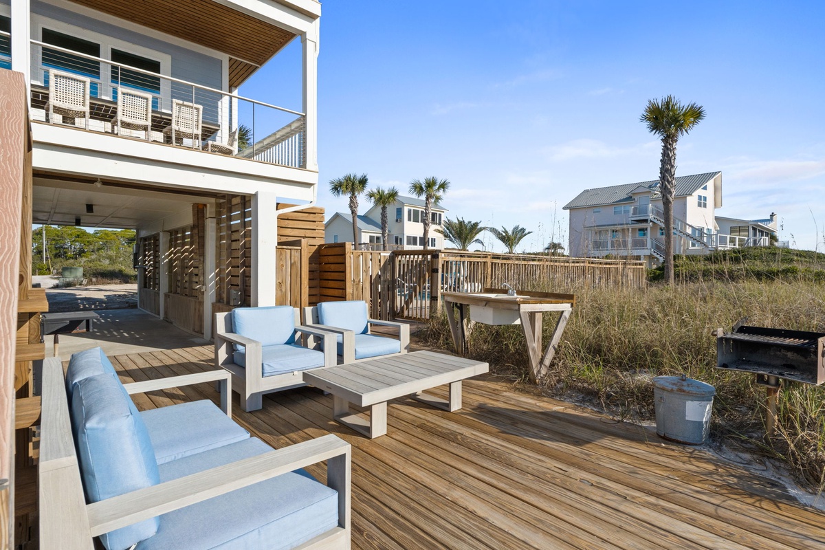 Coastal vacation home exterior featuring covered deck with comfortable seating surrounded by palm trees and nearby beach houses.
