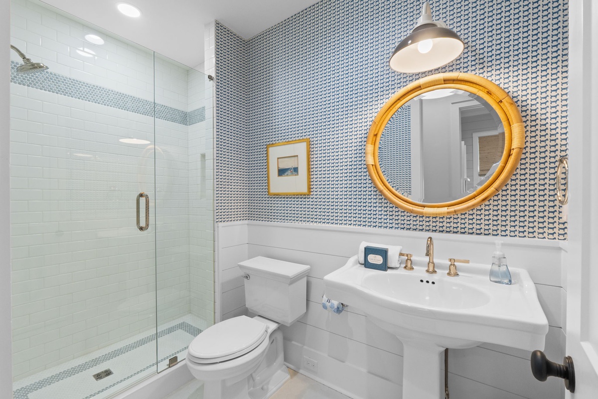 Freshen up in your chic bathroom, featuring a sleek glass shower, elegant tilework, and a bright gold-framed mirror that adds a touch of coastal charm.