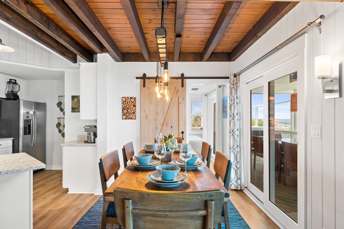 Step into a warm, open-concept space with striking wood-beamed ceilings and a charming barn door that slides open to reveal the heart of your vacation home—a beautifully set dining table ready for meals together and glimpses of the sea beyond.
