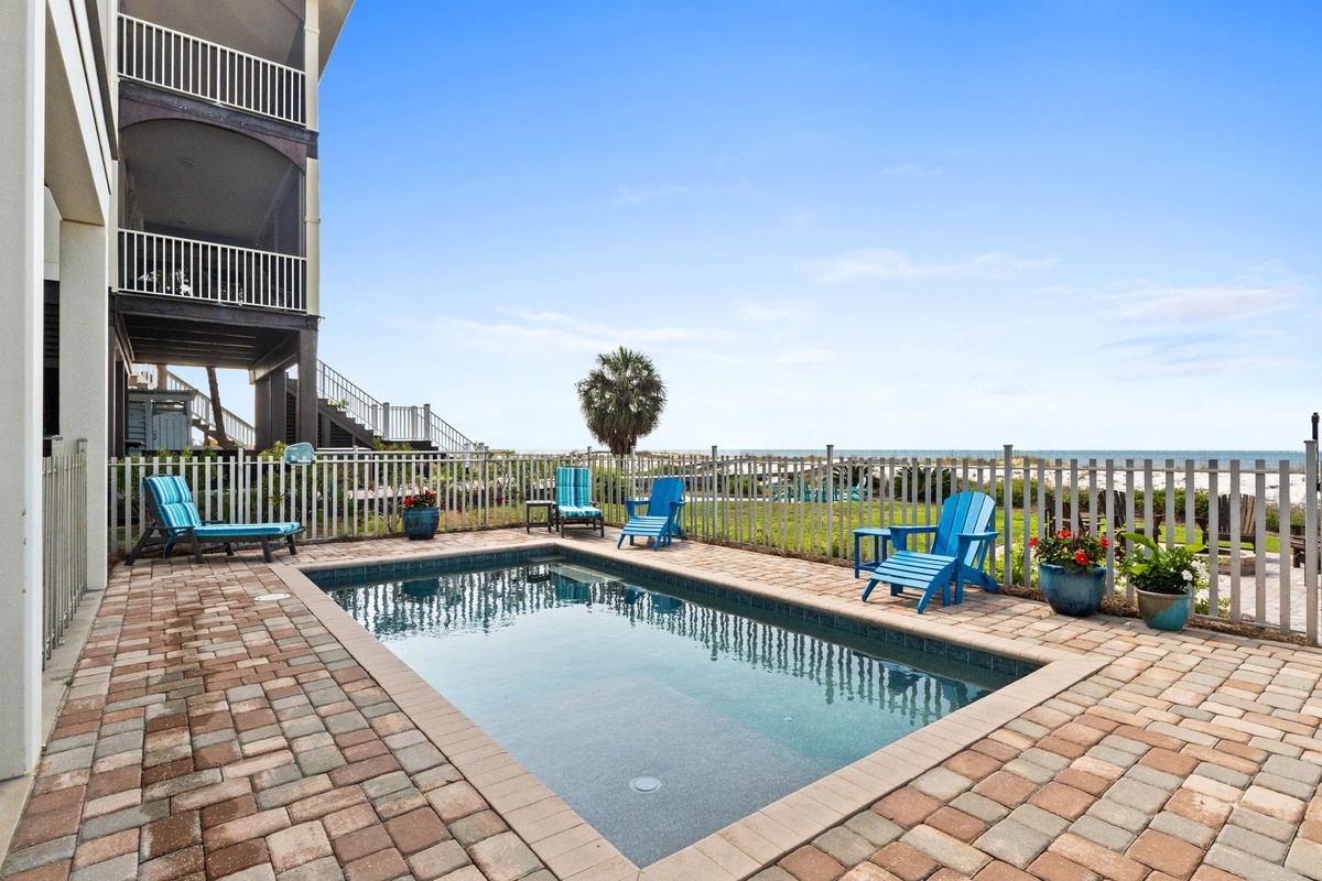 Your private pool awaits, framed by vibrant blue chairs and coastal breezes. Picture yourself lounging poolside with ocean glimpses beyond the fence.
