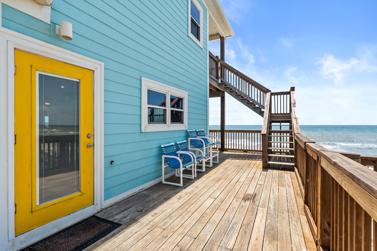 Beachfront property deck with turquoise siding, vibrant yellow door, and direct boardwalk access to the sandy shore and ocean beyond.