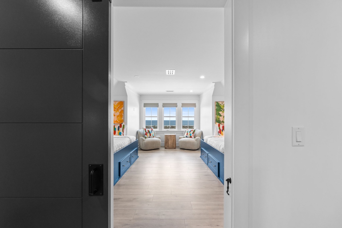 Step into your bright coastal retreat with twin beds, cozy seating, and stunning ocean views framed by three large windows—perfect for relaxing after a day at the beach.