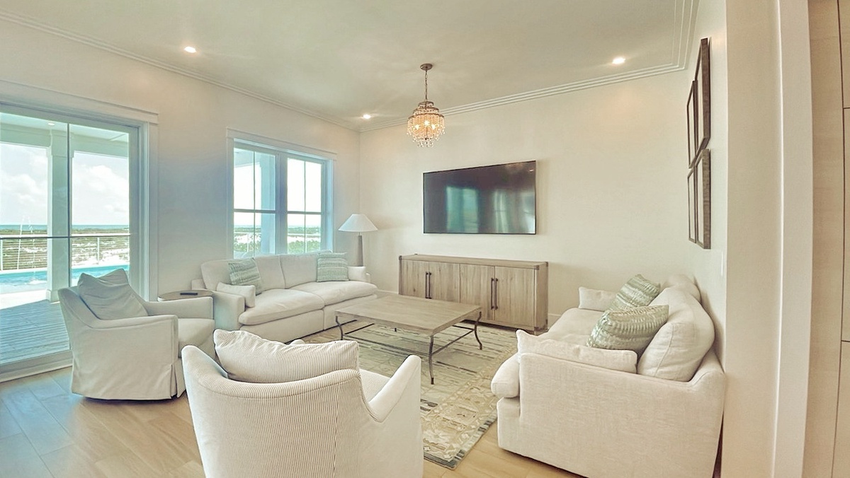 Sink into plush seating and unwind in your bright, airy living room. Large windows frame pool and water views, while soft coastal tones invite relaxation. Your perfect retreat awaits.