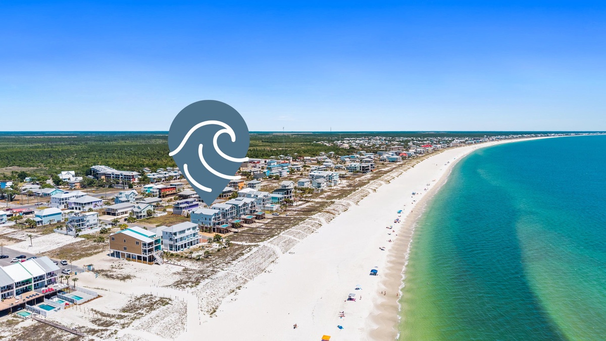 Aerial view captures a pristine beachfront community with white sand coastline stretching along turquoise waters, surrounded by natural landscape and scattered coastal residences.