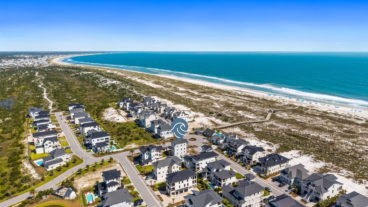 A stunning coastal community positioned along pristine beachfront, bordered by natural dunes and vegetation, with the turquoise ocean stretching to the horizon under clear skies.