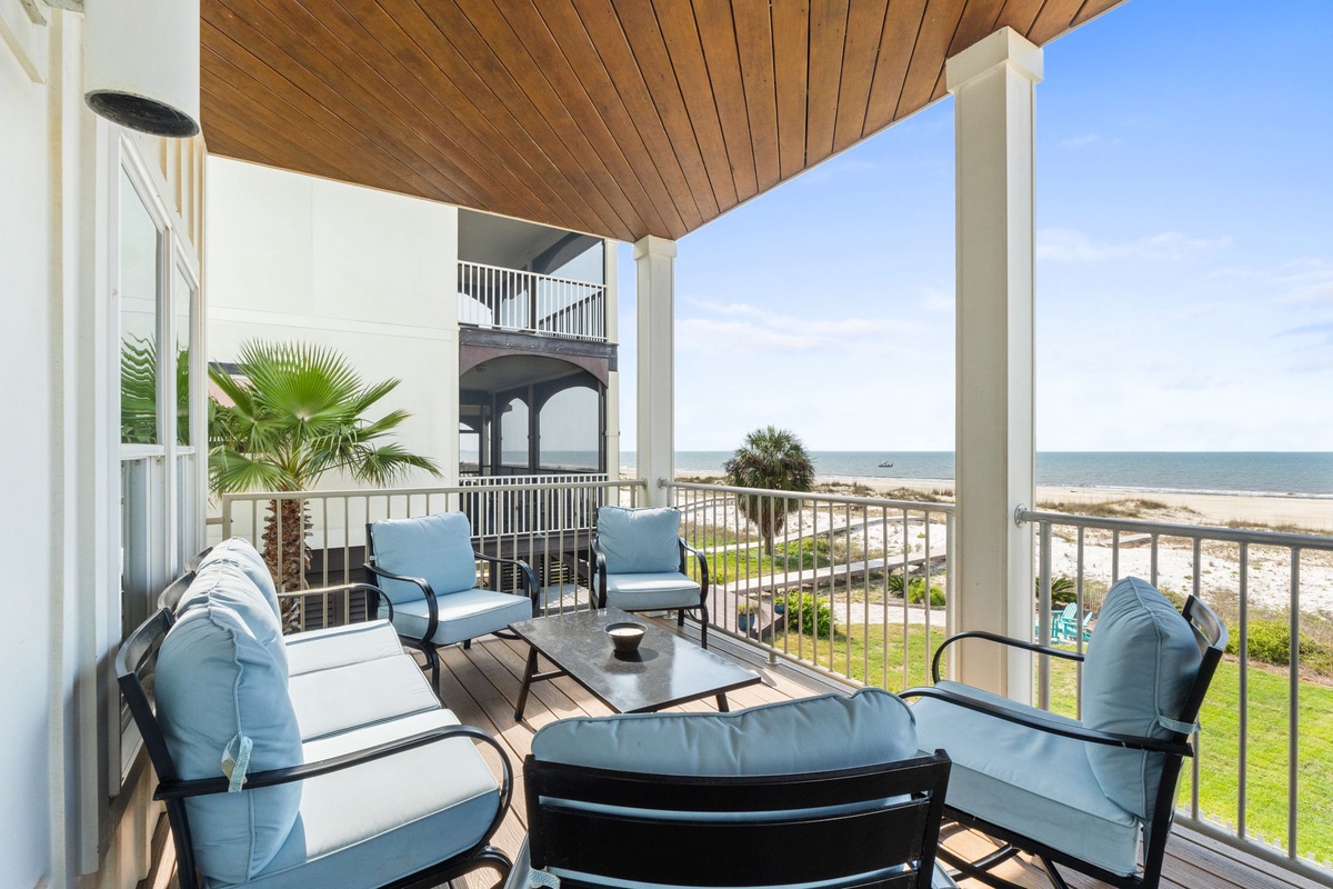 Comfortable waterfront balcony with cushioned seating overlooking the beach. A peaceful spot to unwind while enjoying coastal breezes and views of the ocean, nearby homes, and sandy shoreline.