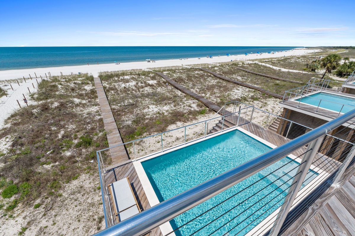 Aerial view of beachfront pools and private boardwalks leading to pristine white sand beach. Crystal-clear turquoise water stretches along sunny coastline with natural dunes and palm trees.