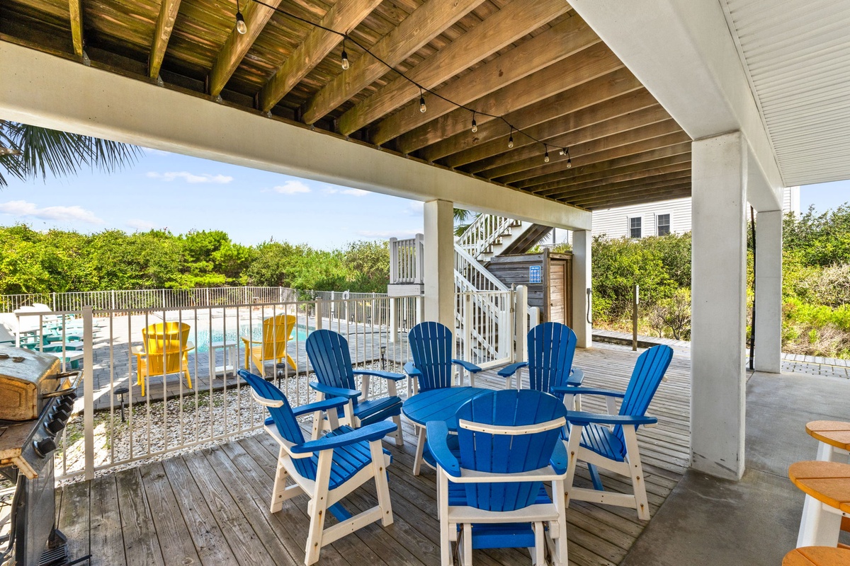 This covered outdoor space is an inviting gathering spot, with colorful Adirondack chairs framing the pool, natural landscaping just beyond, and weathered wood details lending a breezy beach-house feel.