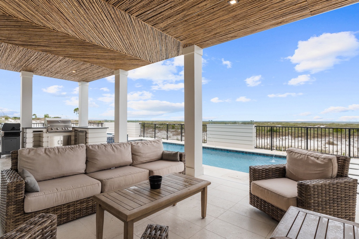 An elevated waterfront lounge space features a natural reed ceiling, BBQ facilities, and modern patio furnishings overlooking a pool and scenic views beyond.