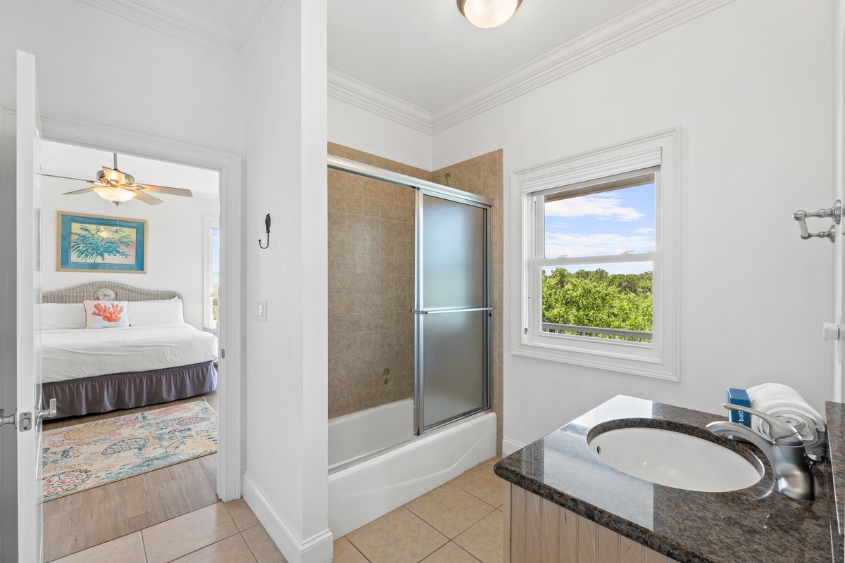 Your bright bathroom features a shower-tub combo with glass doors and a granite-topped vanity. The window frames lush treetop views that bring the outdoors in.