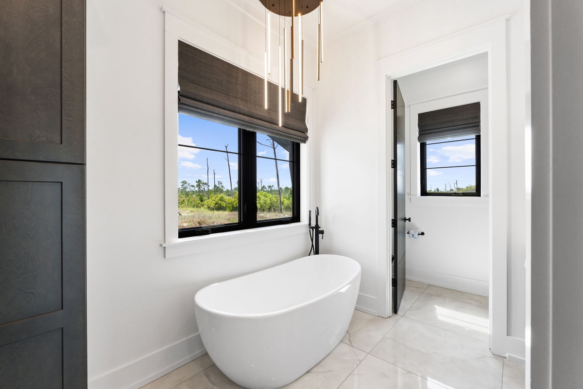 Slip into the freestanding soaking tub beneath elegant pendant lighting, where natural light streams through large windows framing peaceful landscape views—your personal sanctuary for ultimate relaxation.