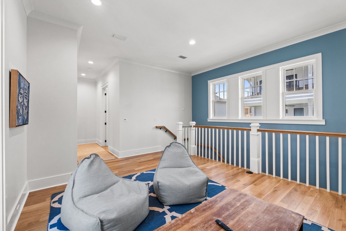 A spacious upstairs landing with warm hardwood floors and soothing blue accent walls. Sink into the comfortable bean bag seating and enjoy natural light streaming through charming windows overlooking the property.