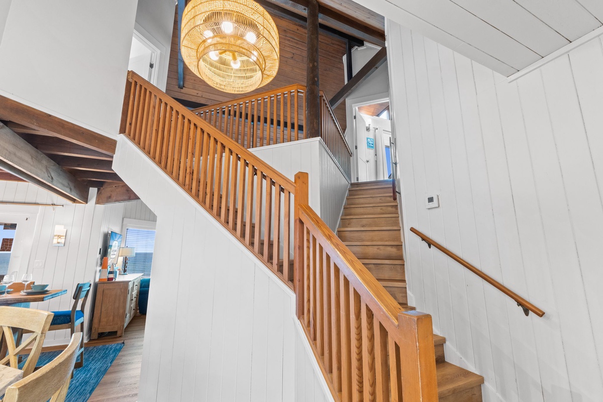 Natural wood stairs wind upward through open, sunlit spaces, drawing you into a vacation home where warmth and coastal ease blend under a statement woven chandelier.