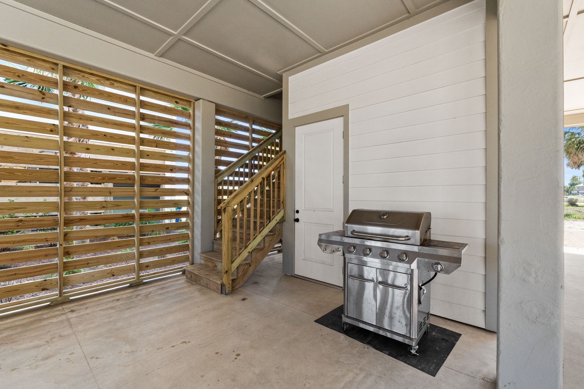A covered ground-level area features a stainless steel grill ready for your outdoor cooking. Wooden slat privacy screening and a staircase leading up complete this functional space beneath the home.
