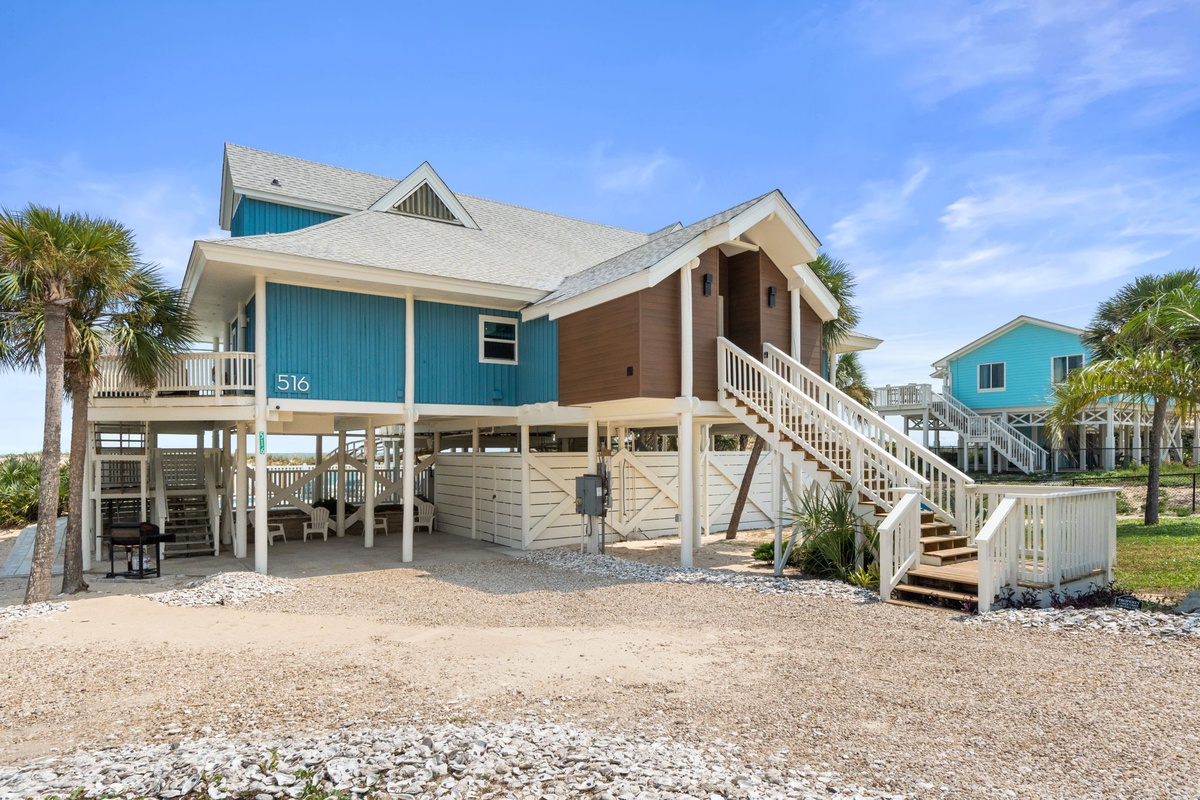 Cheerful beach house elevated on stilts with tropical palms and coastal neighbors nearby. The elevated design and staircase entrance signal proximity to sandy shores and beachside living.