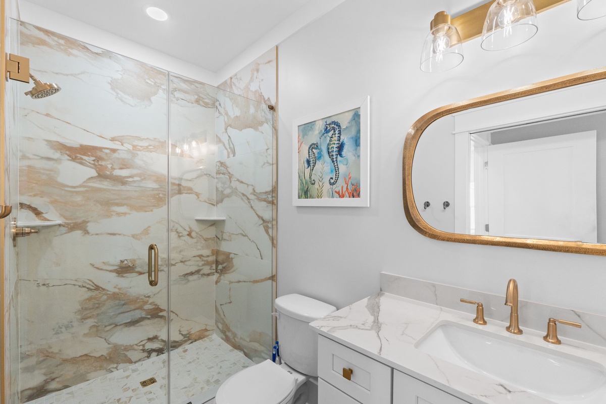 Your spa-like bathroom features a stunning walk-in shower with elegant marble-patterned walls, gleaming gold fixtures, a polished vanity, and coastal-inspired art to help you unwind in style.
