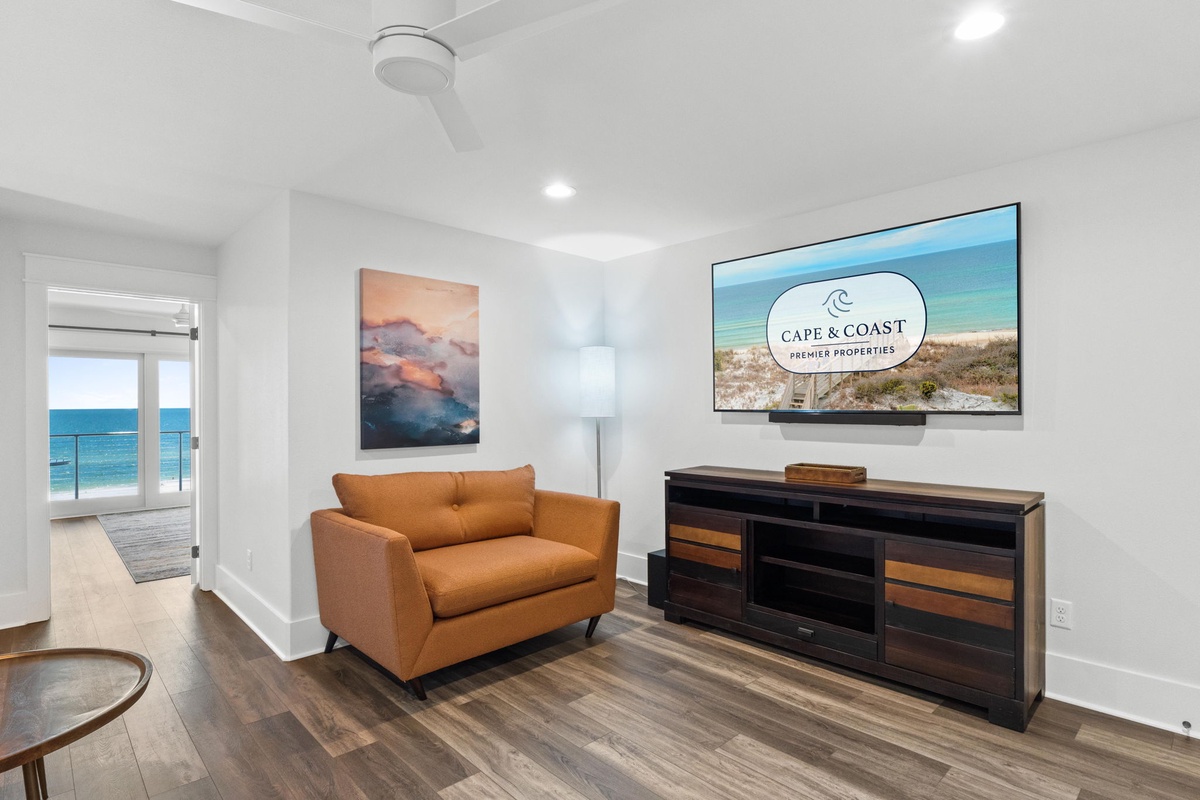 Relax in your cozy living room with coastal artwork and a large TV. The open doorway invites you to the sunlit balcony with ocean views.