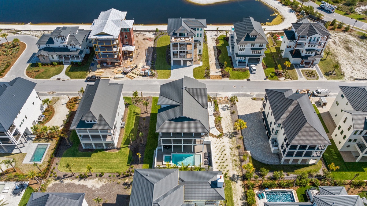Aerial view of a coastal vacation community featuring modern beachfront homes with private pools and landscaped yards, situated along calm waterfront streets just steps from the beach.