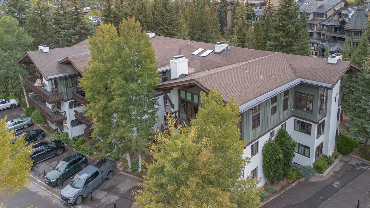 Aerial view of the property nestled among tall evergreens and autumn foliage, with convenient parking and nearby buildings visible in the surrounding mountain resort area.