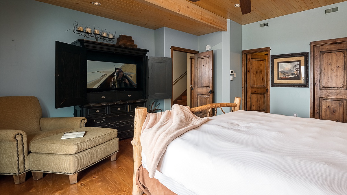Unwind in your spacious bedroom suite, featuring a plush king bed, entertainment center, and cozy sitting area beneath warm wood-beam ceilings. The perfect blend of mountain charm and modern comfort.