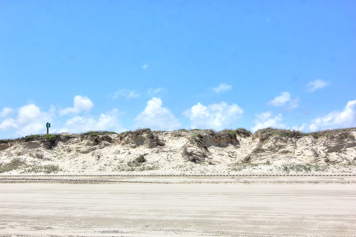 Natural sand dunes stretch along the coastline under bright blue skies with scattered white clouds.