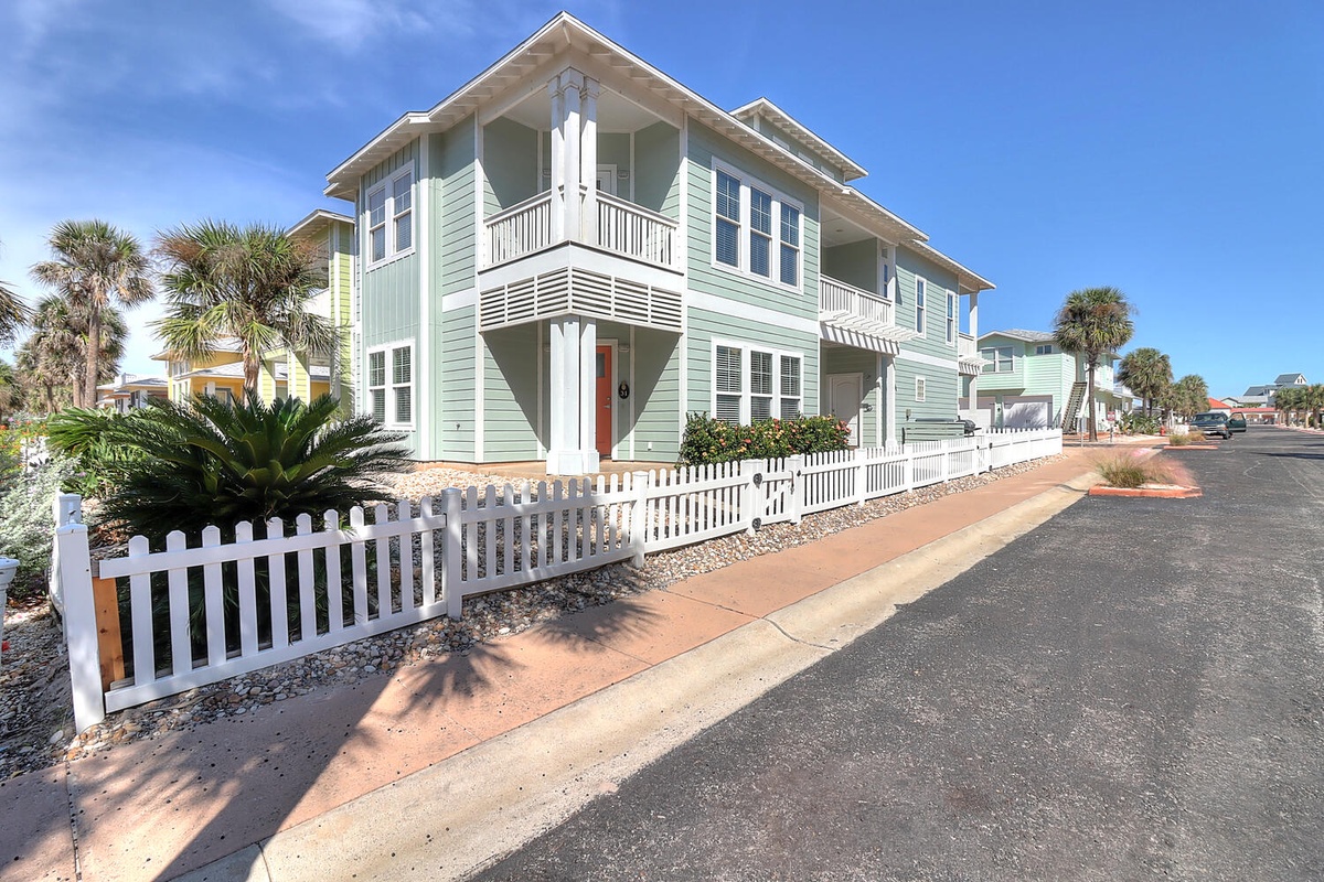 Two-story coastal rental home featuring mint green siding, white picket fencing, and tropical palm landscaping in a charming beachside neighborhood.