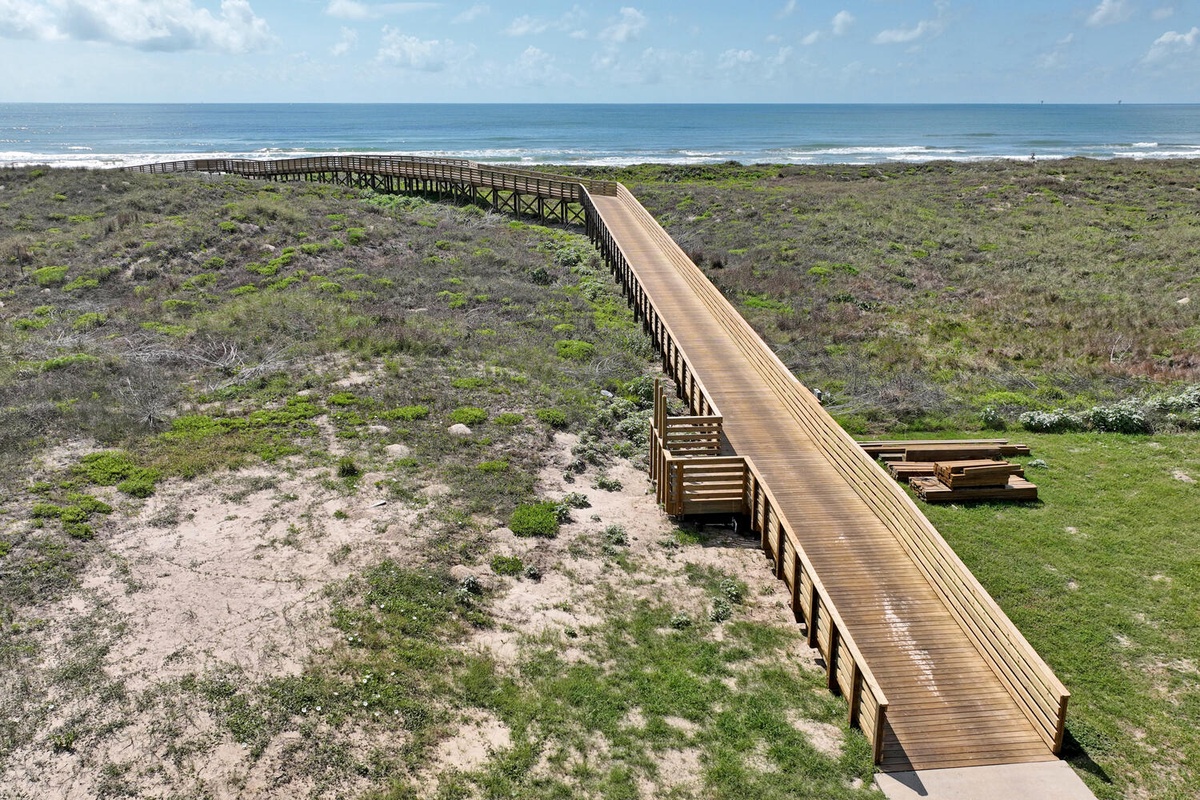 Wooden boardwalk winds through coastal dunes to pristine beach with rolling waves under expansive blue sky.