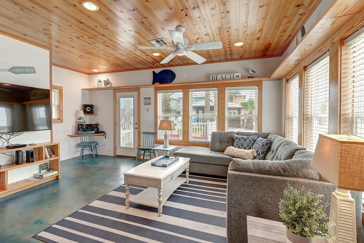 Relax in your bright beach-themed living room with comfy seating, warm wood ceilings, and peaceful neighborhood views through large windows.