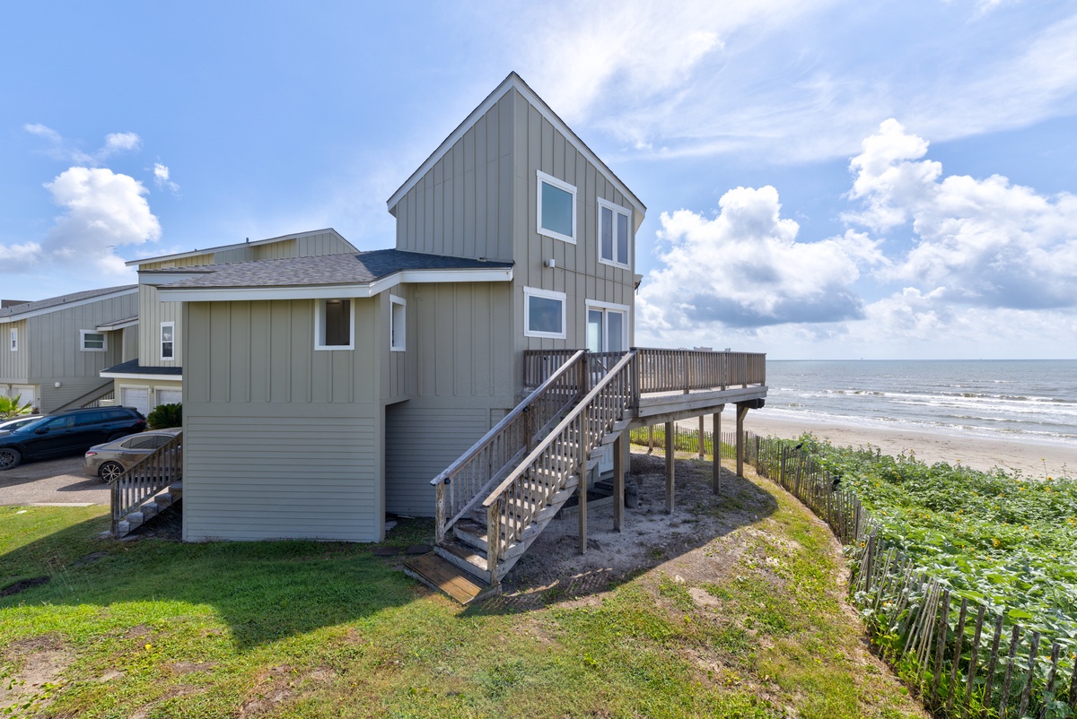 Beachfront vacation home featuring a two-story design with direct ocean access via elevated deck. The property sits on a coastal dune with sandy beach and rolling waves stretching along the shoreline.