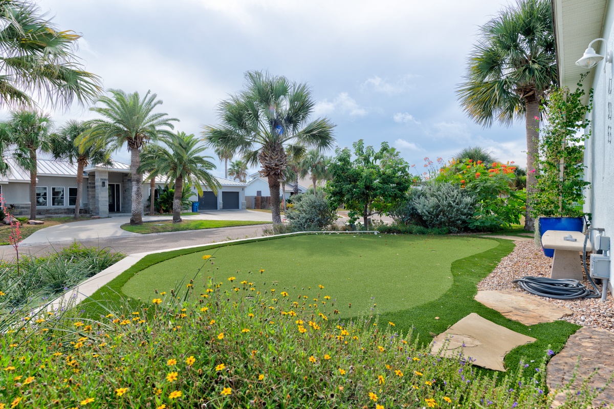 The property features a lush tropical garden with mature palm trees, manicured lawn, and vibrant flowering beds set beneath a peaceful coastal sky.