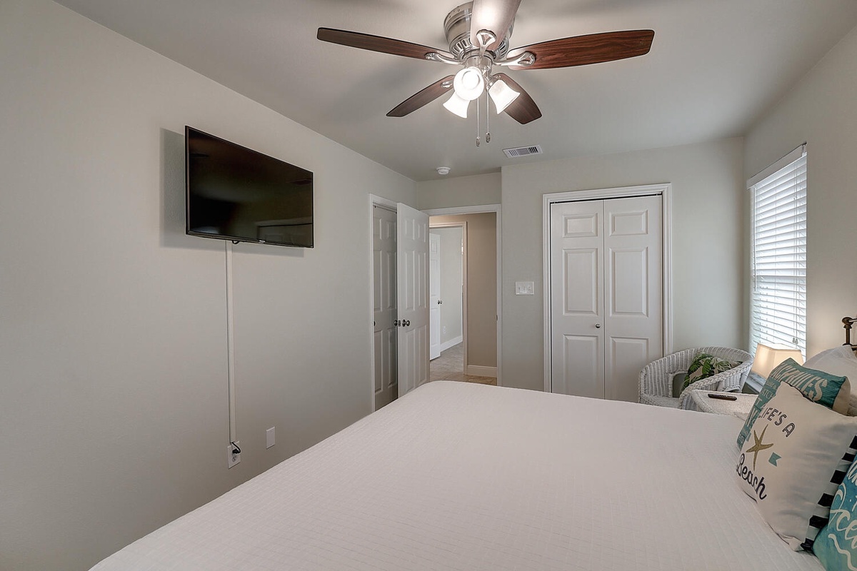 Relax in your spacious bedroom with crisp white linens, ceiling fan, wall-mounted TV, and natural light streaming through the windows.