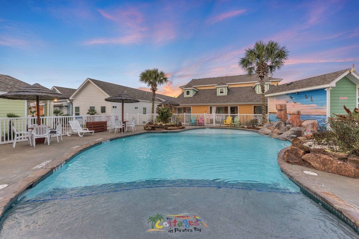 Colorful beach houses surround a sparkling pool area with decorative rock features under a beautiful sunset sky.