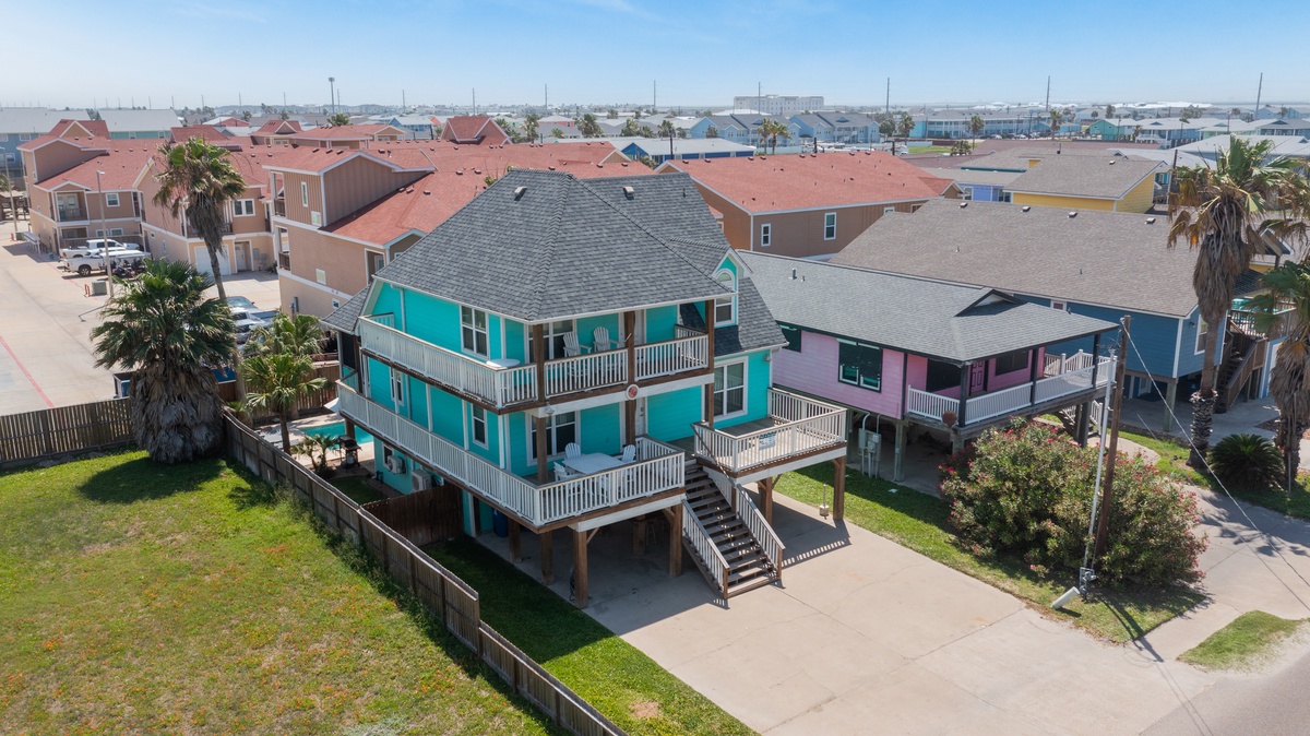 Bright turquoise beach house with spacious decks sits in a vibrant coastal neighborhood, surrounded by colorful homes and swaying palm trees.