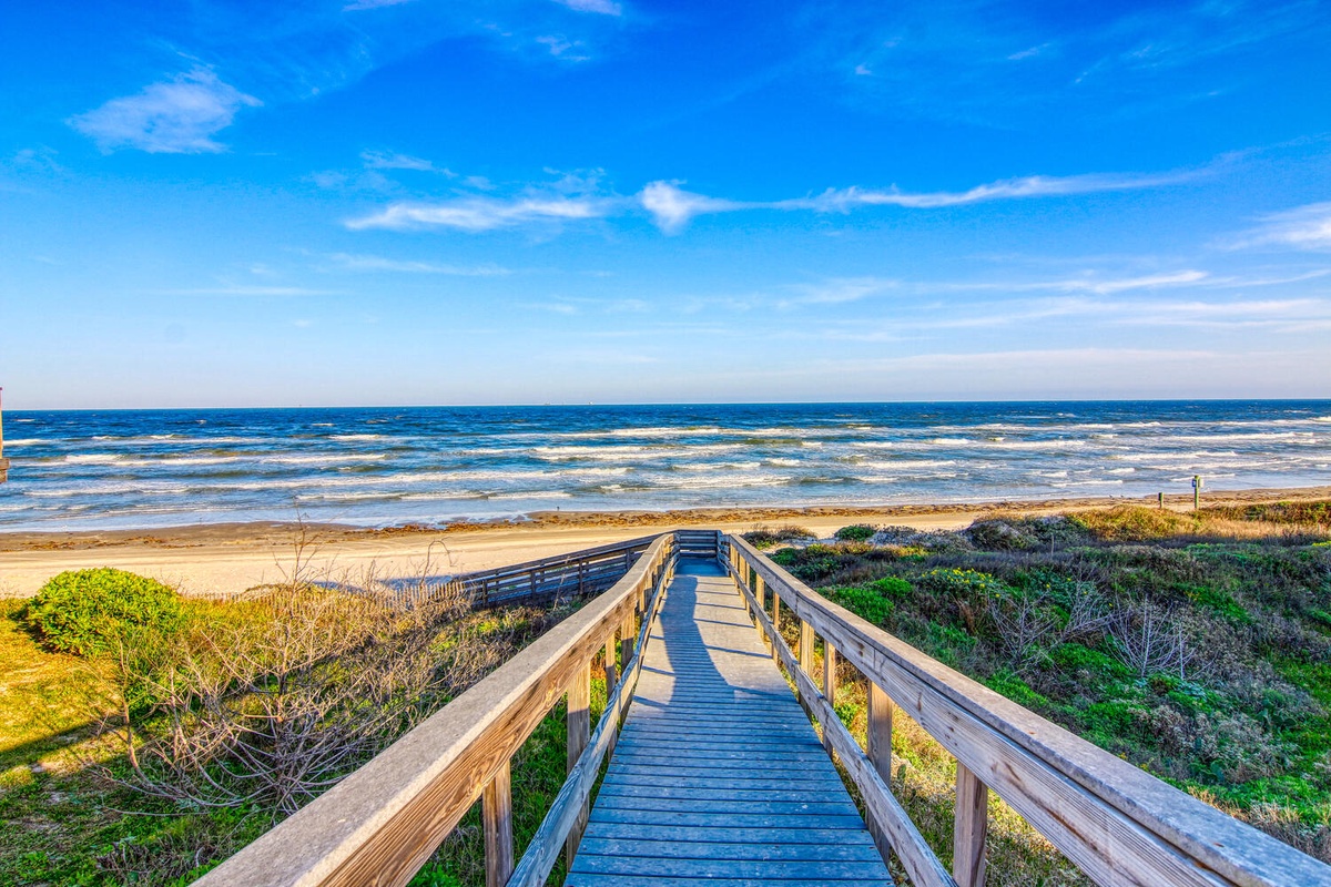 Wooden boardwalk leads through coastal dunes to pristine sandy beach with rolling ocean waves under brilliant blue skies.
