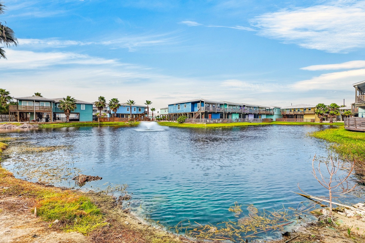 A tranquil waterfront community with colorful buildings surrounding a scenic pond, complete with palm trees and a fountain centerpiece.