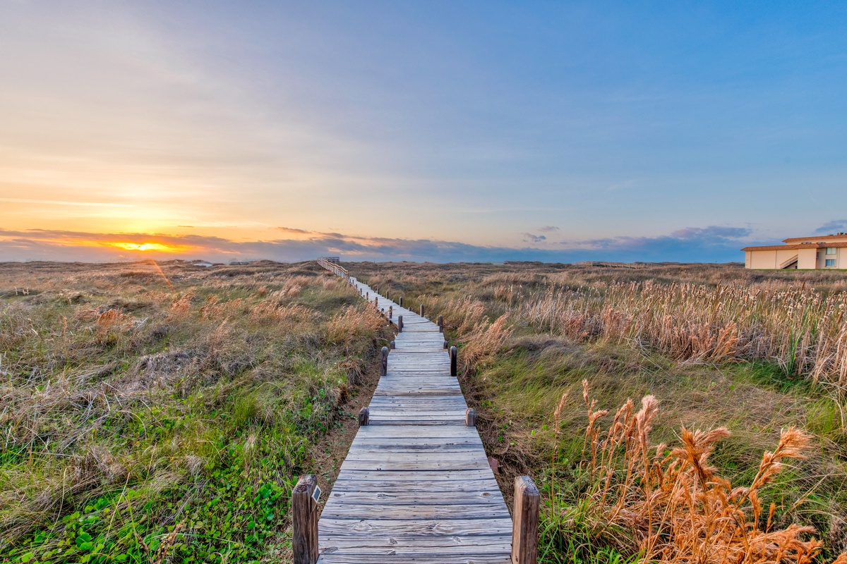 A wooden boardwalk winds through coastal dunes toward a golden sunset, with the property visible in the distance.