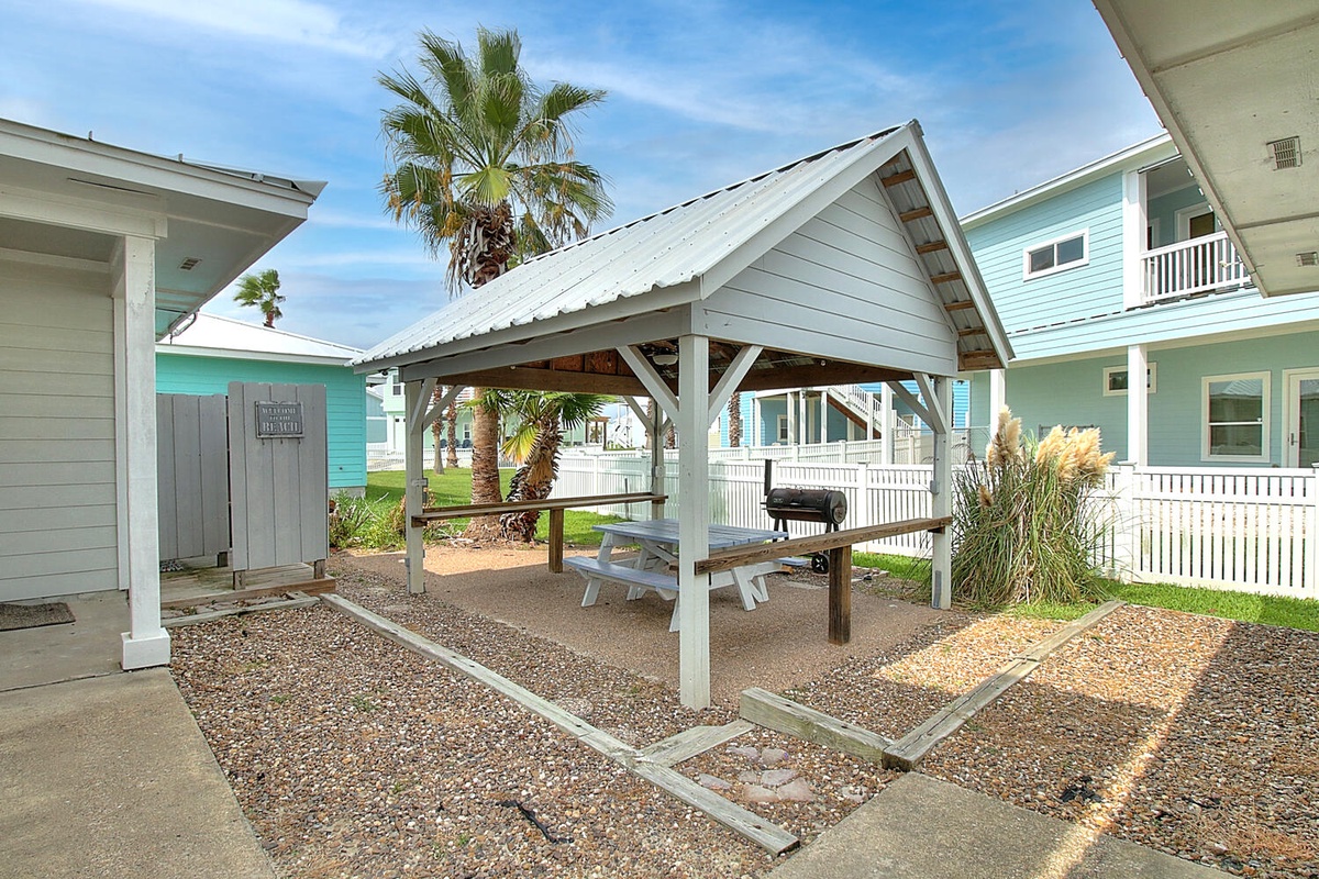 Covered BBQ pavilion with picnic tables set among coastal vacation rentals and swaying palms.