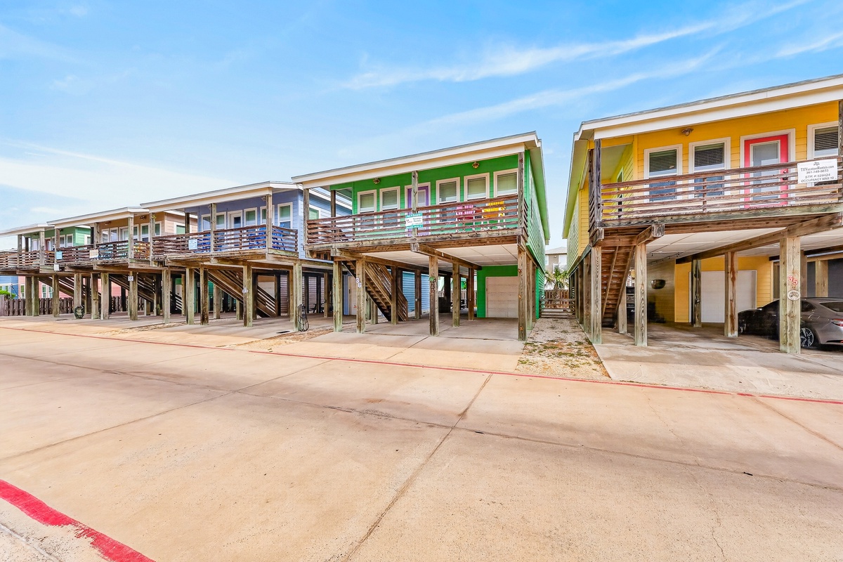 Vibrant stilted beach homes with colorful facades and covered parking below. Each unit features balconies with modern railings and easy access to the sandy shores nearby.