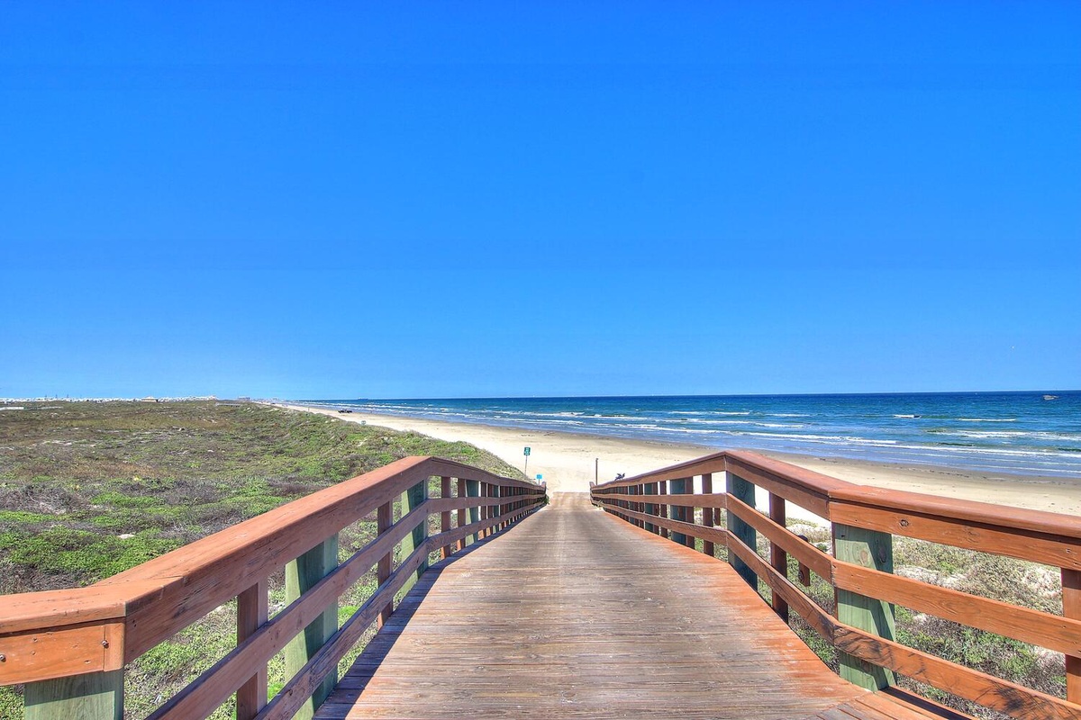 A wooden boardwalk leads to pristine beach with rolling waves under clear blue skies.