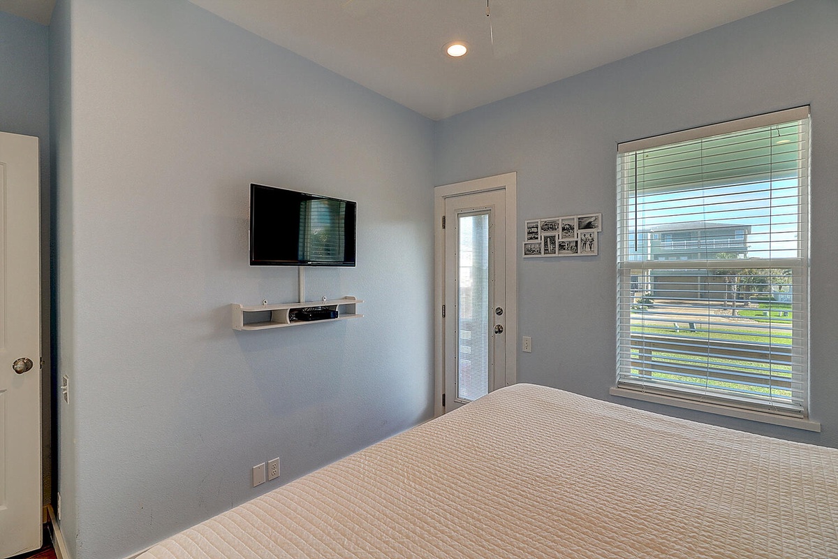 Your tranquil bedroom features soft blue walls, a wall-mounted TV for relaxing evenings, and large windows with blinds for privacy.