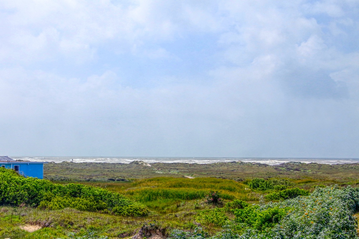 Coastal dunes and natural vegetation stretch toward the ocean, with blue sky and distant waves creating a serene seaside setting.