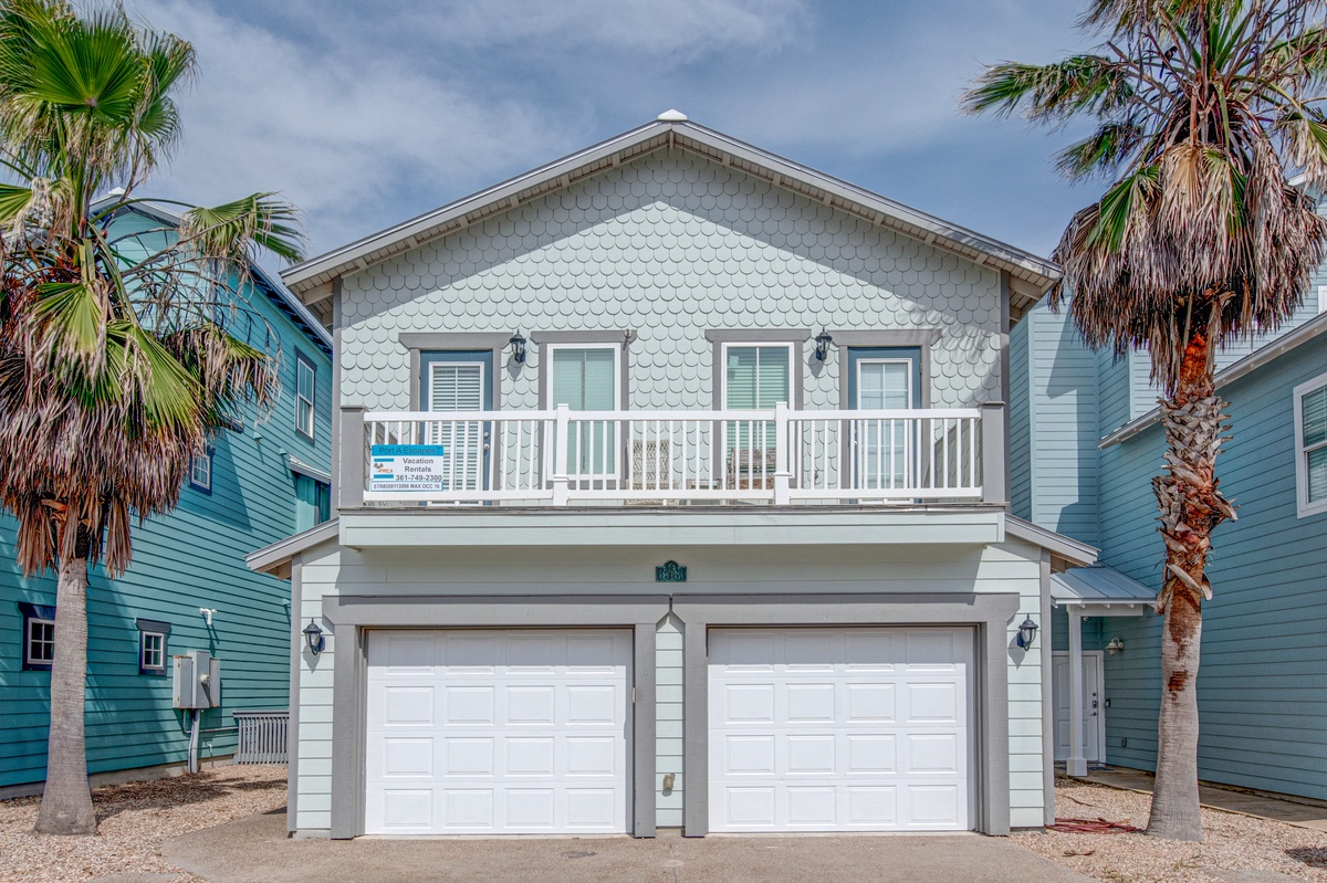 A charming coastal rental home with distinctive shingle siding and tropical palm trees, featuring a welcoming balcony and convenient two-car garage in a vibrant beach neighborhood.