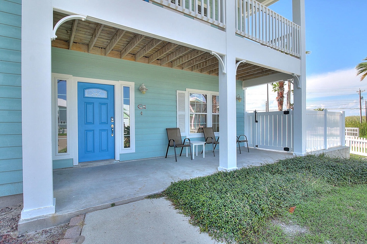 Bright coastal beach house with covered porch, outdoor seating, and cheerful blue entrance door welcoming guests to their seaside getaway.