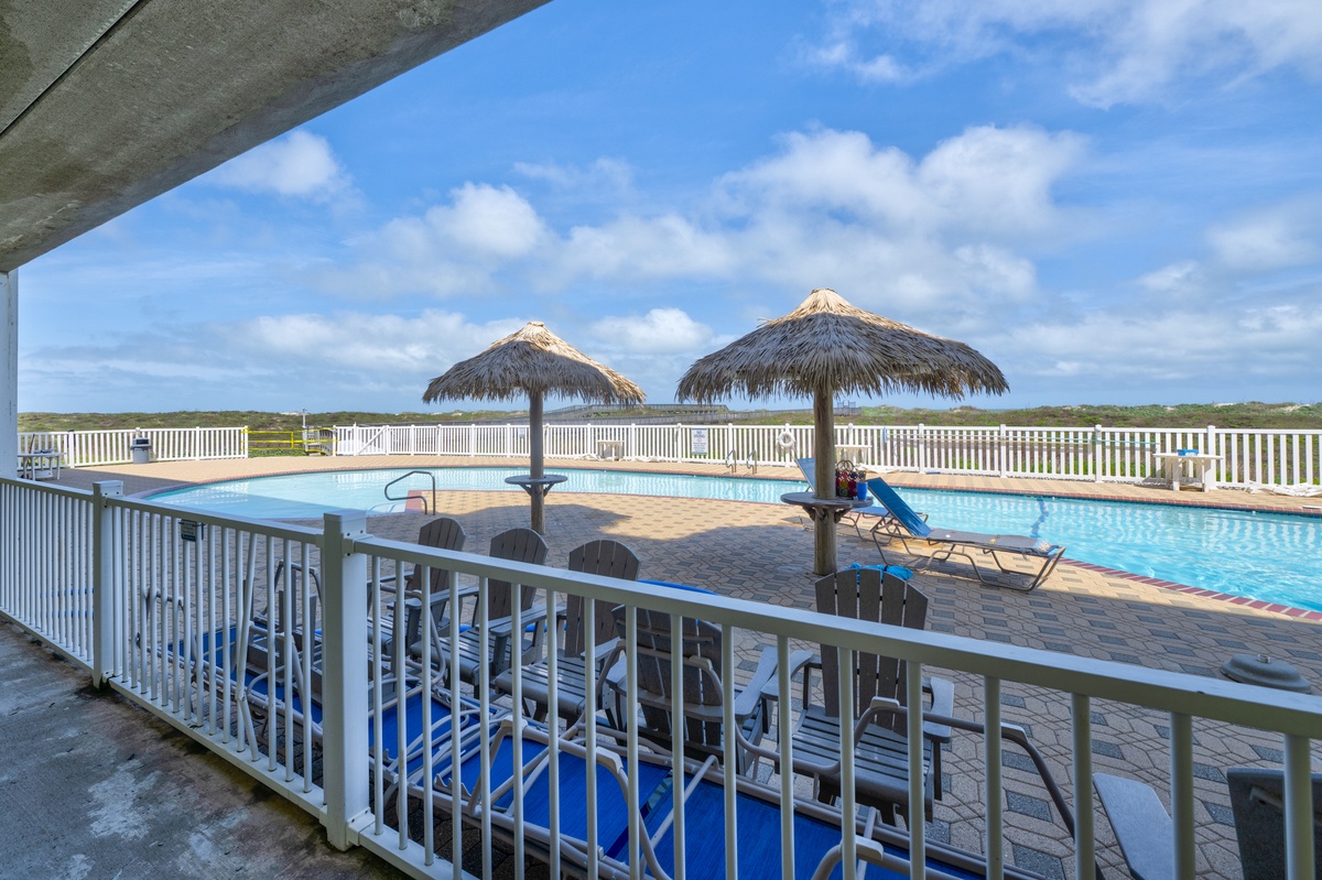 Expansive pool area features thatched palapa umbrellas and loungers, bordered by white fencing overlooking open natural landscape beyond the property.