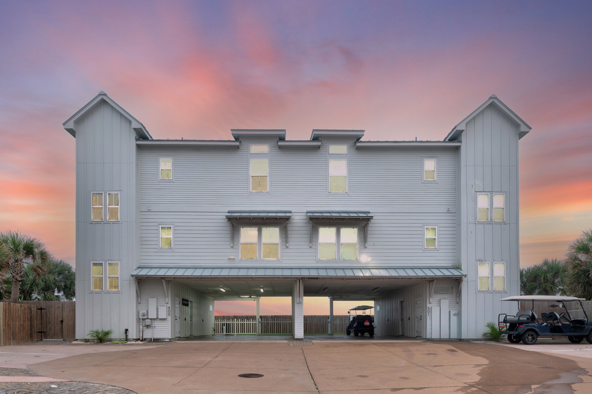 Modern coastal property featuring gray siding and warm lighting against dramatic sunset skies.