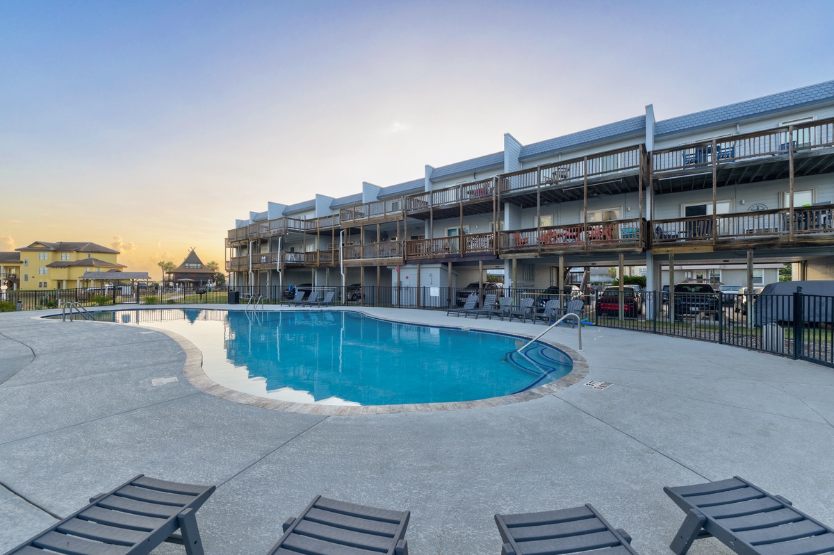Multi-level property building with private balconies surrounding a sparkling outdoor pool, photographed during golden hour with ample sun loungers on the poolside deck.