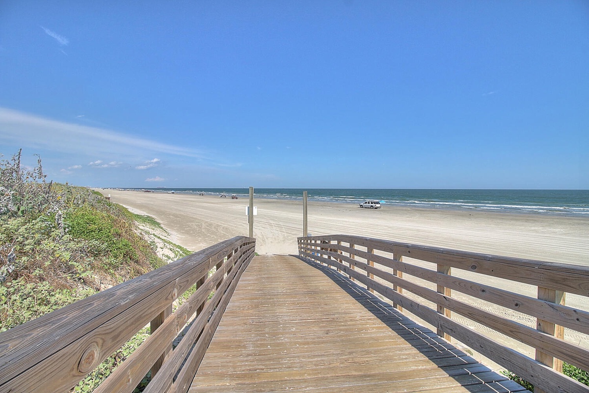 Wooden boardwalk leads directly to pristine sandy beach with ocean waves and clear blue skies stretching endlessly ahead.