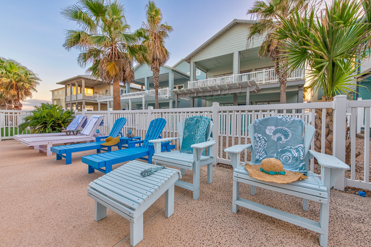 Beachfront relaxation area with comfortable lounge chairs surrounded by palm trees and coastal property buildings.
