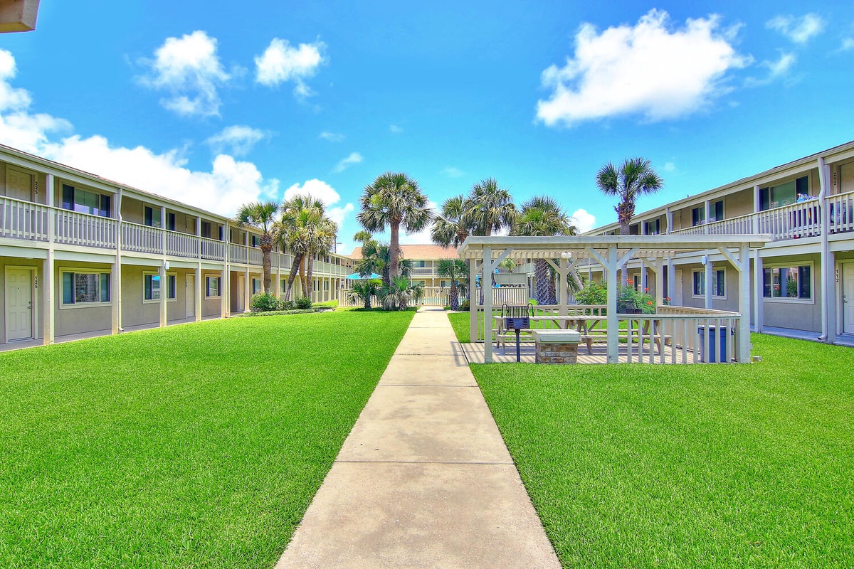 Tropical property courtyard with lush green lawn, palm trees, and covered pavilion under bright blue skies.