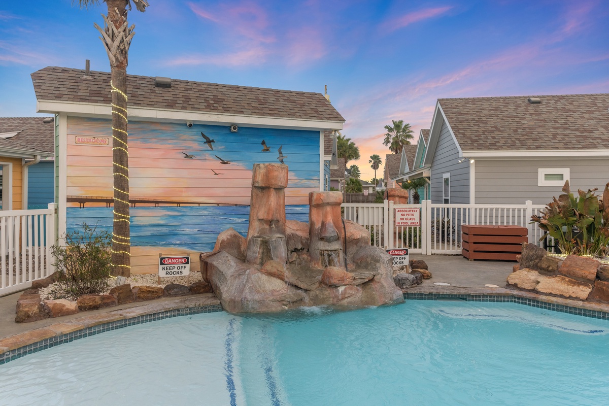 Tropical swimming pool with rock waterfall feature surrounded by colorful beach-themed cottages and palm trees.